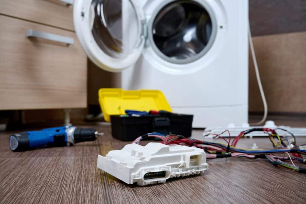 Réparation Machine à Laver : Module Électronique et Outils Réparation de machine à laver avec module électronique, fils colorés, perceuse et boîte à outils sur le sol.
